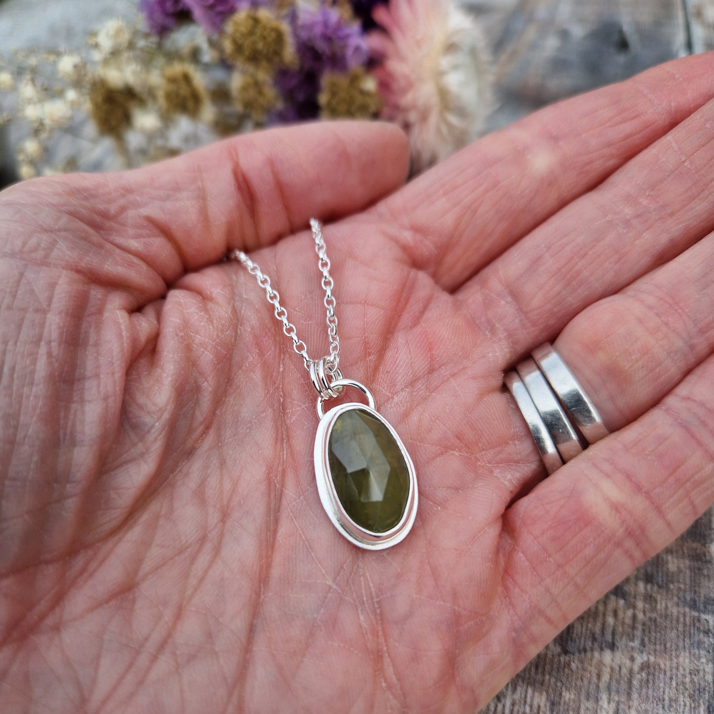 Green sapphire oval gemstone with diamond cut texture, set in simple silver bezel, attached to silver chain via two small circle links.