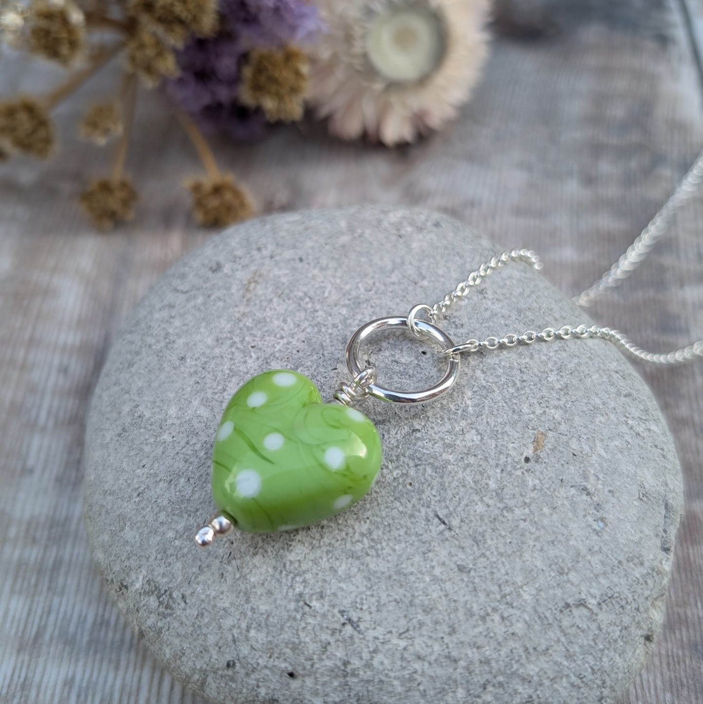 A close-up of a vibrant green heart-shaped Lampwork glass pendant with white polka dots, hanging on a recycled sterling silver necklace, placed on a textured stone background with dried flowers.