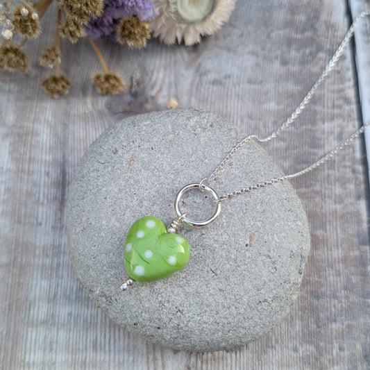 A close-up of a vibrant green heart-shaped Lampwork glass pendant with white polka dots, hanging on a recycled sterling silver necklace, placed on a textured stone background with dried flowers.