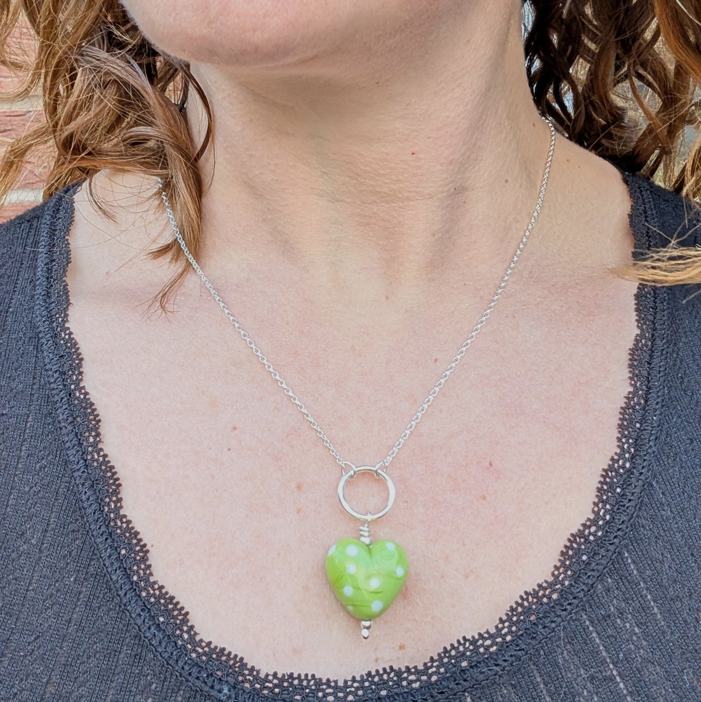 A model wearing a recycled sterling silver necklace featuring a green polka-dot Lampwork glass heart pendant, styled against a dark top.