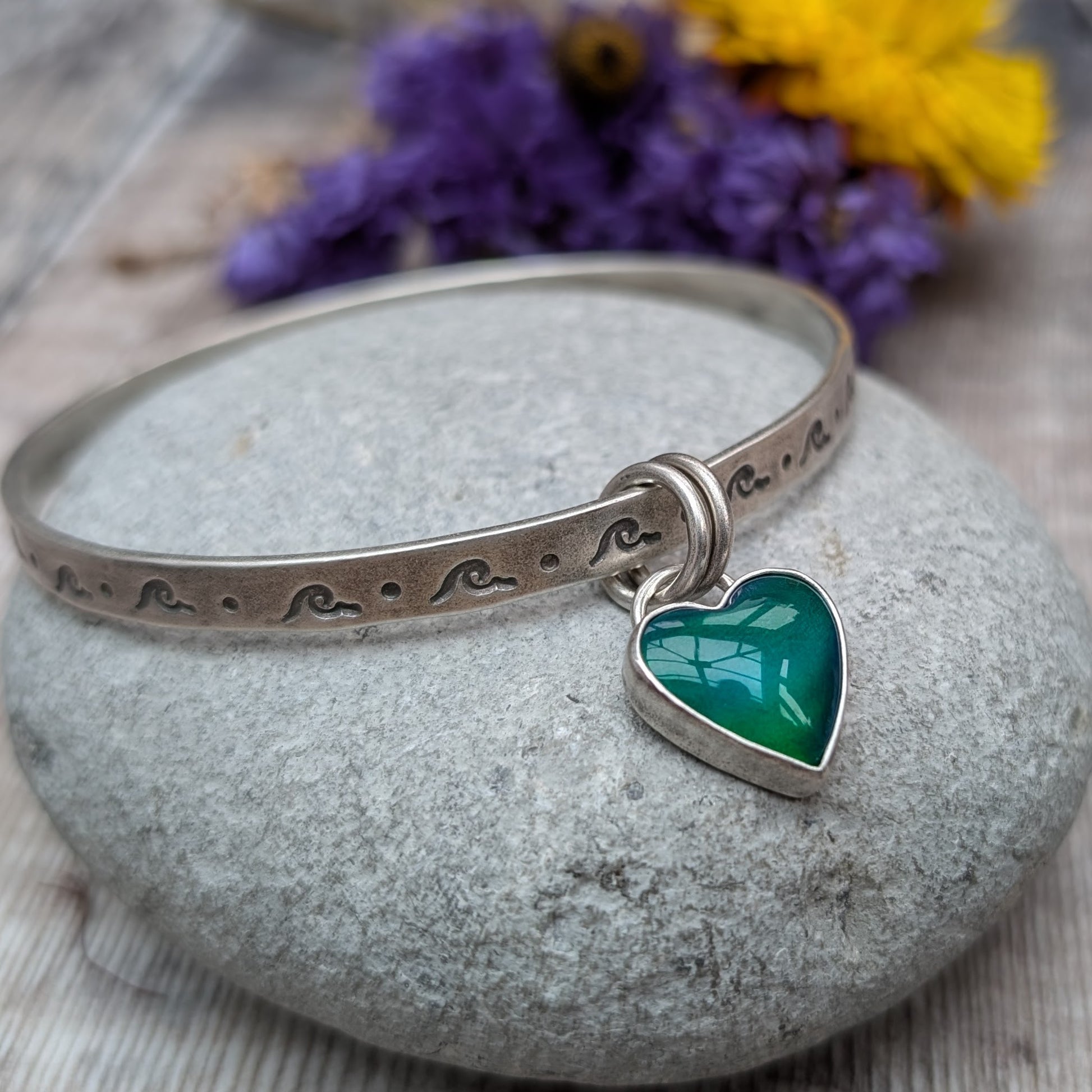 A silver bangle with wave-shaped etchings lies on a smooth, grey stone. The bangle features a heart-shaped charm with a green and blue design, attached by two small rings.