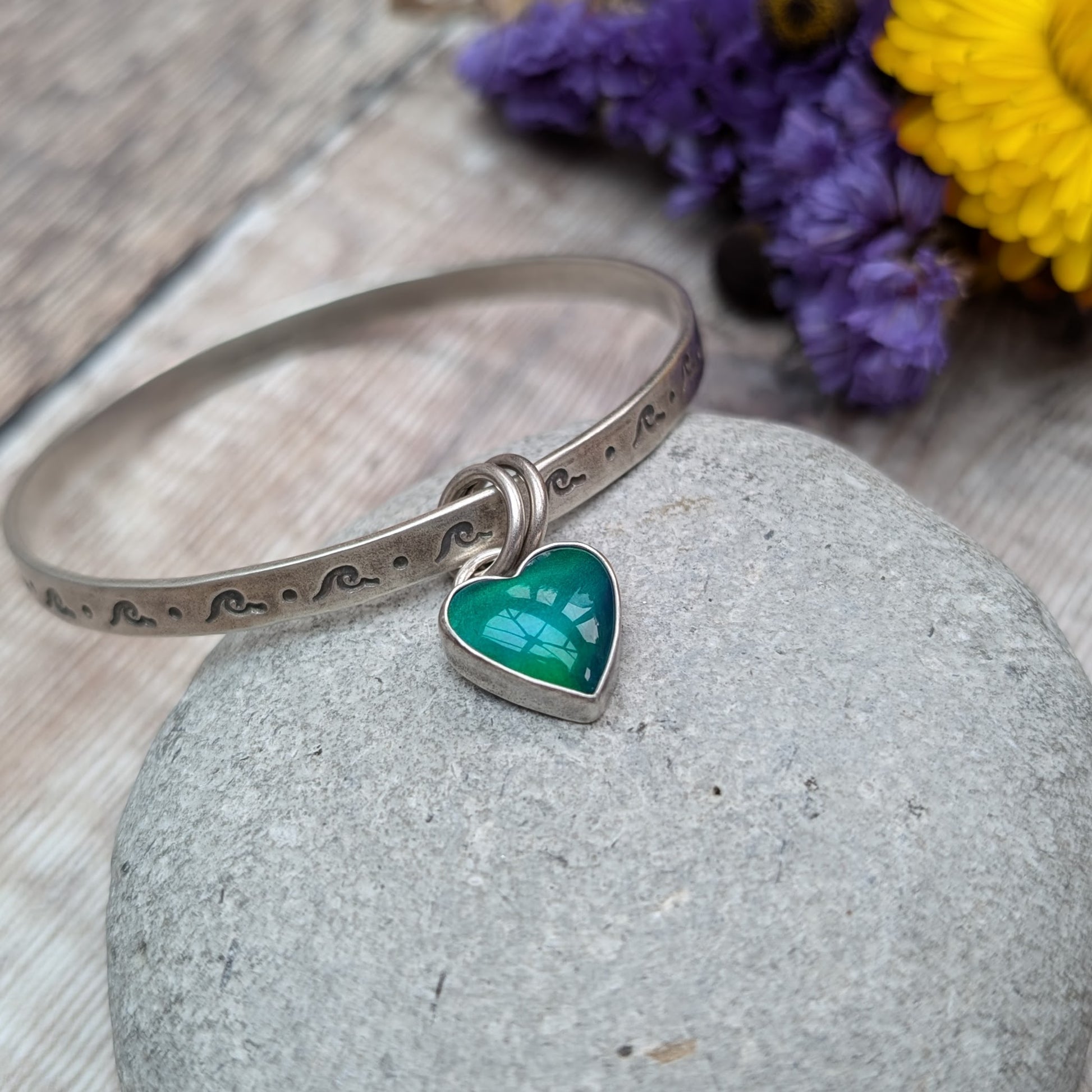 A silver bangle with wave-shaped etchings lies on a smooth, grey stone. The bangle features a heart-shaped charm with a green and blue design, attached by two small rings.