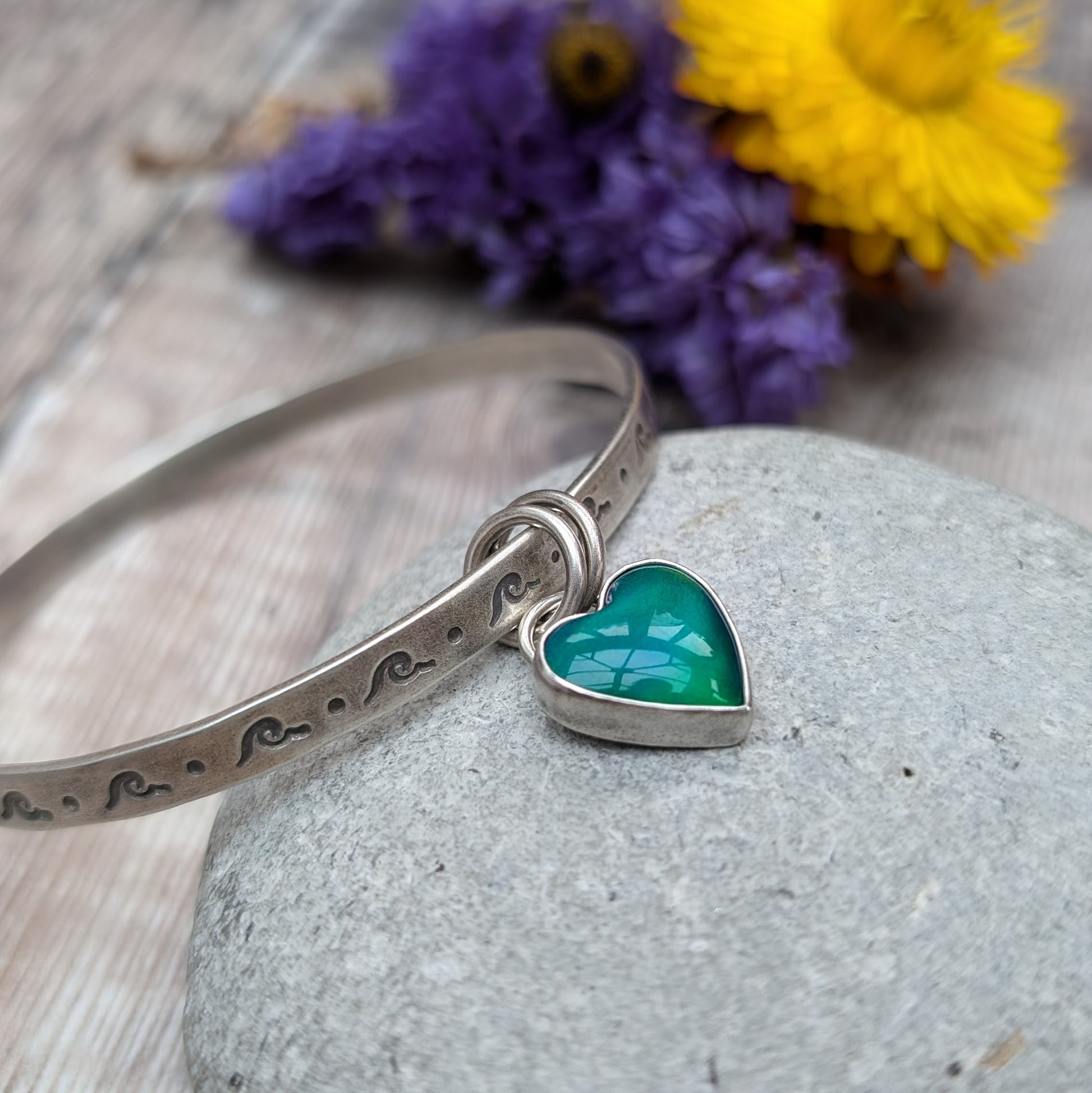 A silver bangle with wave-shaped etchings lies on a smooth, grey stone. The bangle features a heart-shaped charm with a green and blue design, attached by two small rings.