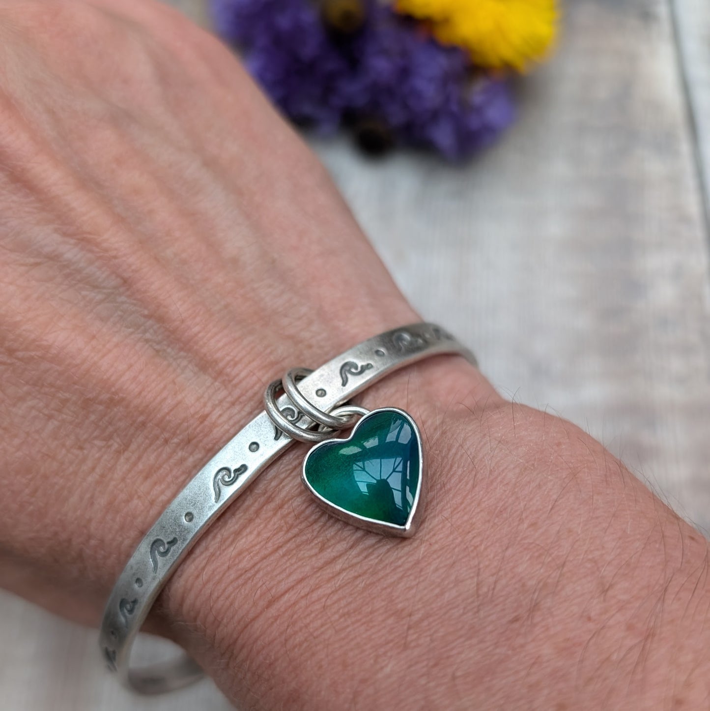 A close-up of a person's wrist wearing a silver bangle with wave-shaped etchings. The bangle features a heart-shaped charm with a green and blue design, attached by two small rings.
