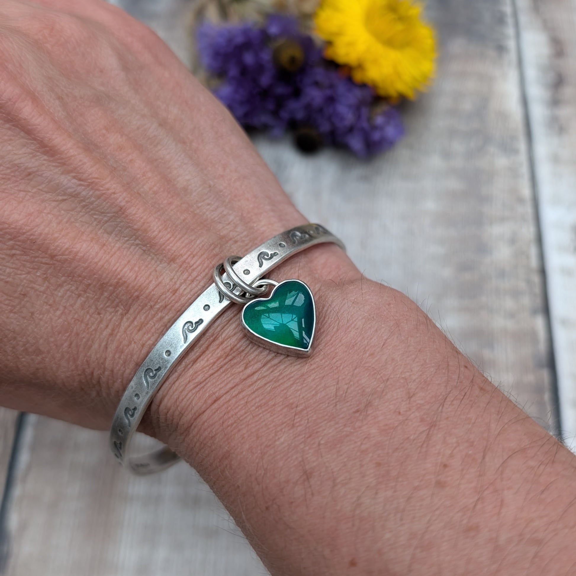 A close-up of a person's wrist wearing a silver bangle with wave-shaped etchings. The bangle features a heart-shaped charm with a green and blue design, attached by two small rings.