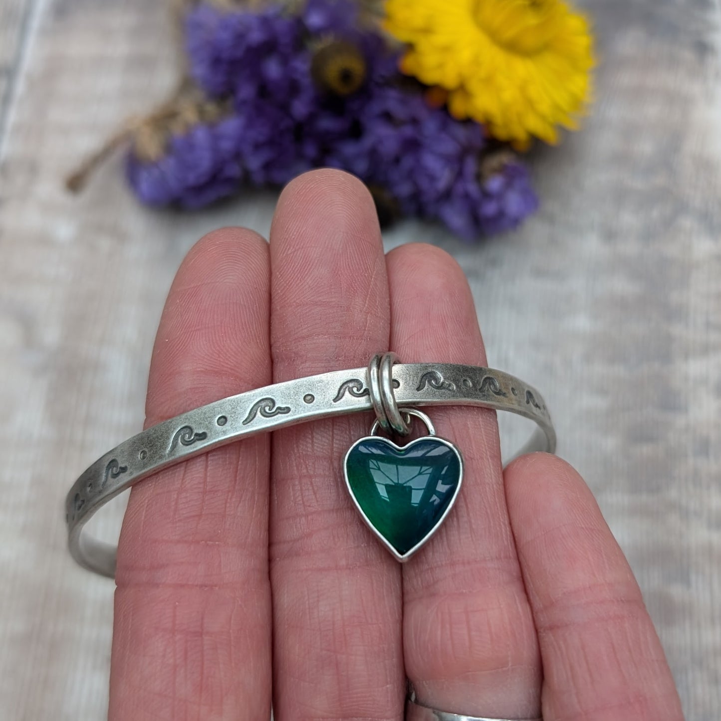 A close-up of a person's wrist wearing a silver bangle with wave-shaped etchings. The bangle features a heart-shaped charm with a green and blue design, attached by two small rings.