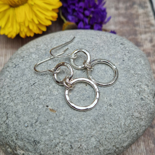 Sterling Silver two circle drop earrings. Each earring has one smaller silver open circle attached to silver earring wire. Dropping from the first open circle via two small silver hoops is a larger silver open circle. Each circle has slightly hammered texture, the largest circle measuring approximately 15mm diameter. Total drop from earlobe approximately 35mm providing lots of movement.