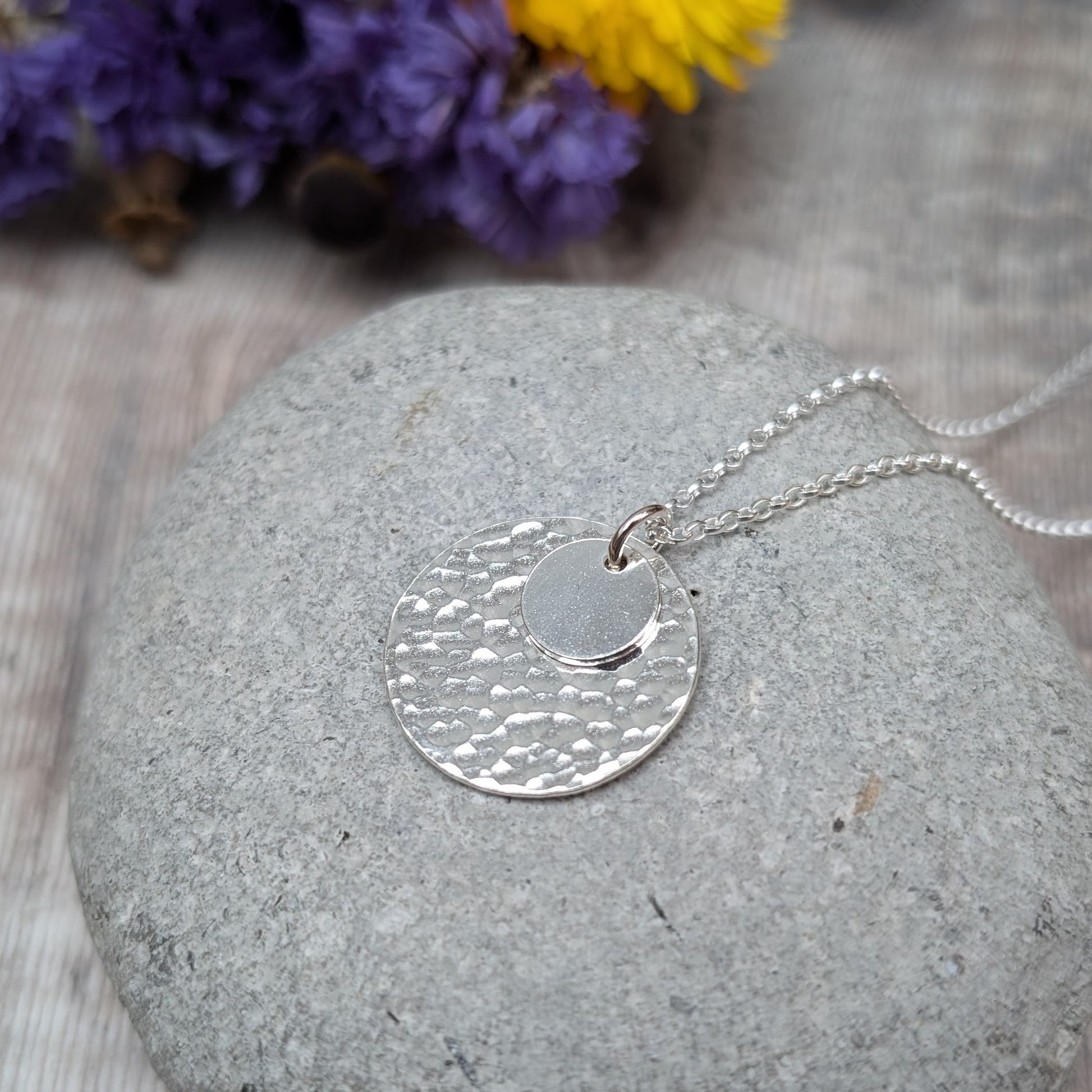 Handmade silver necklace featuring two hammered discs, one small and one large, displayed on a smooth grey stone with blurred purple and yellow flowers in the background.