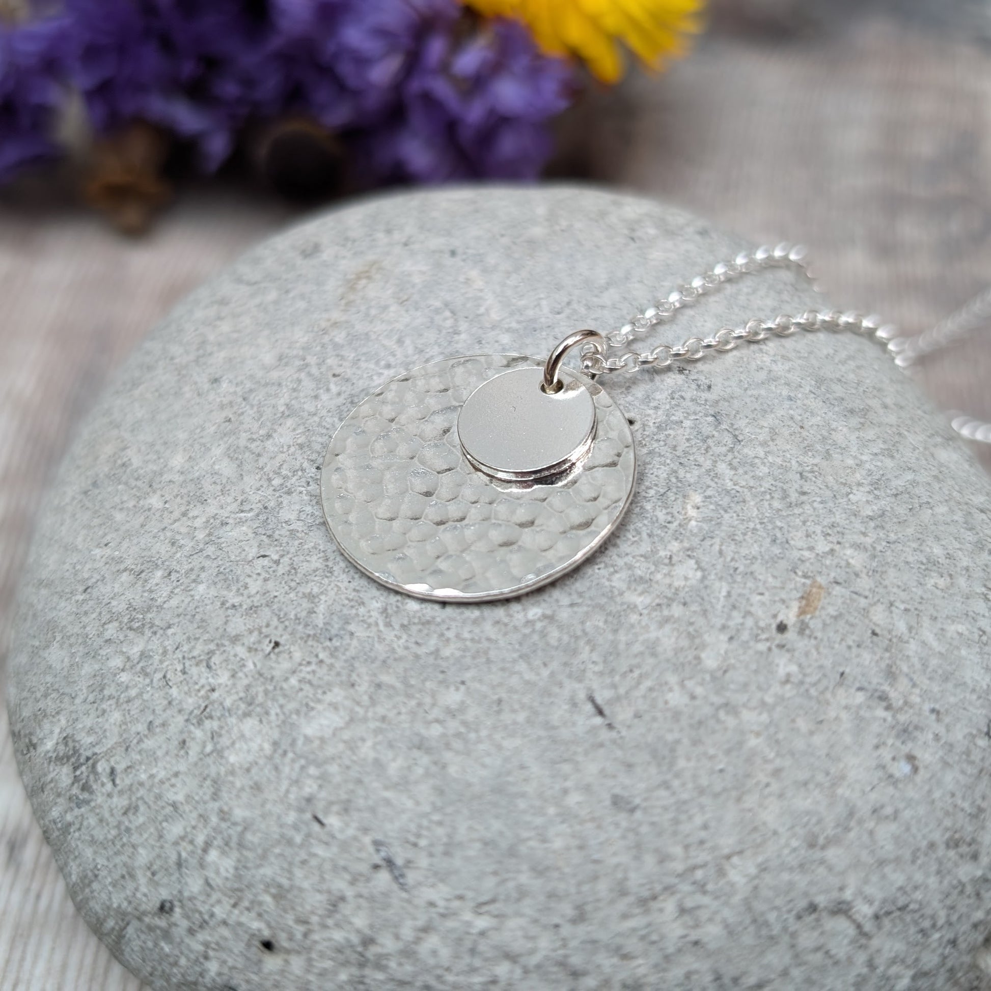 Handmade silver necklace featuring two hammered discs, one small and one large, displayed on a smooth grey stone with blurred purple and yellow flowers in the background.