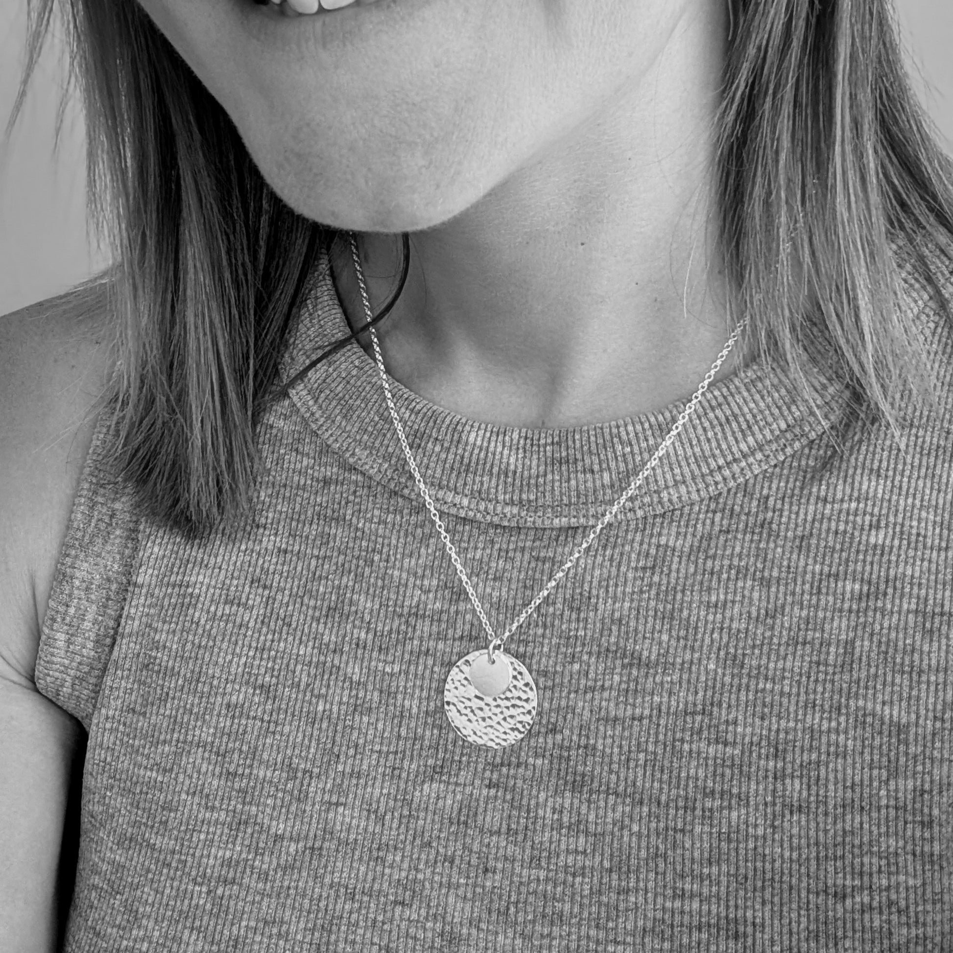 Black and white image of a woman wearing a handmade silver necklace with two hammered discs, one small and one large, against a ribbed sleeveless top.