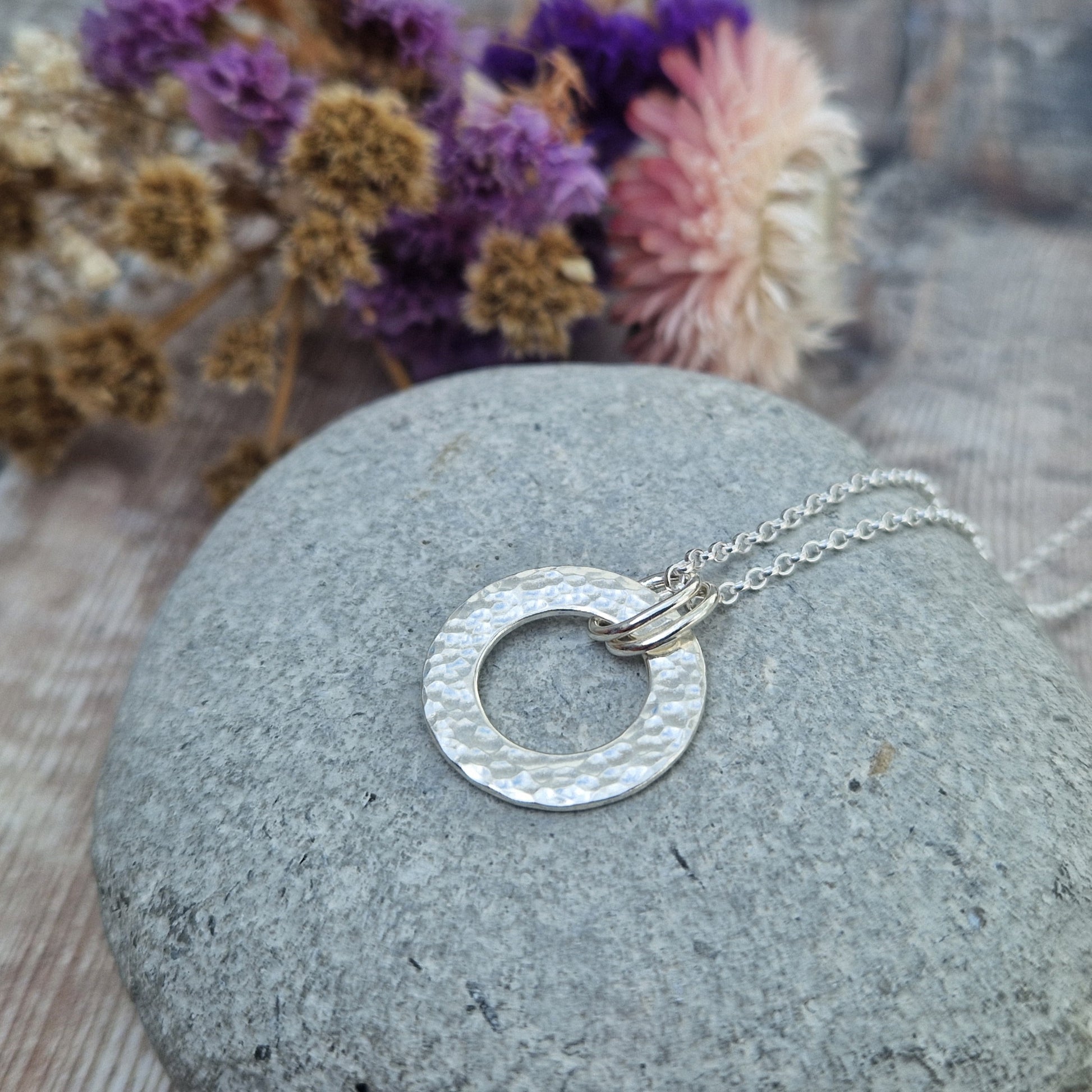 Handmade silver hammered circle necklace laid on a single smooth grey stone, with soft purple and beige dried flowers in the background.
