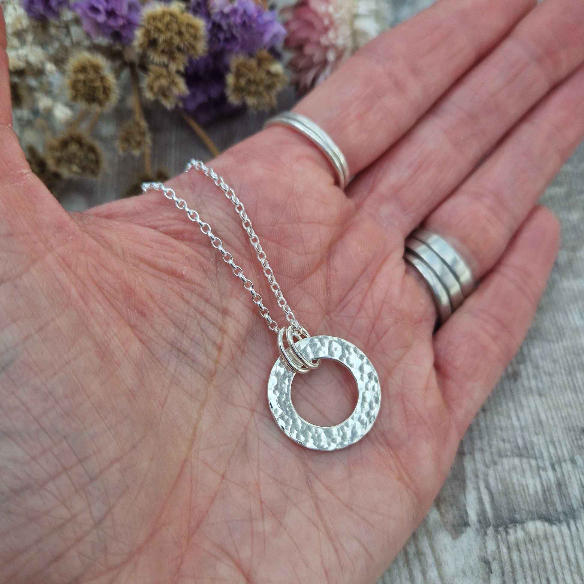 Handmade silver hammered circle necklace held in the palm of a hand, with several silver rings worn on the fingers, and dried flowers blurred in the background.