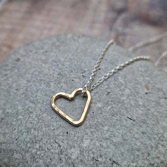 A close-up view of a hammered gold open heart pendant hanging from a silver chain, placed on a smooth stone background.