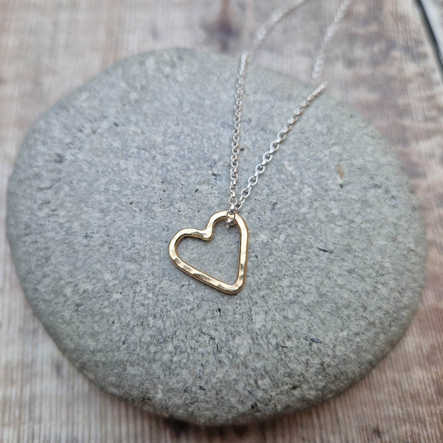 A close-up view of a hammered gold open heart pendant hanging from a silver chain, placed on a smooth stone background.