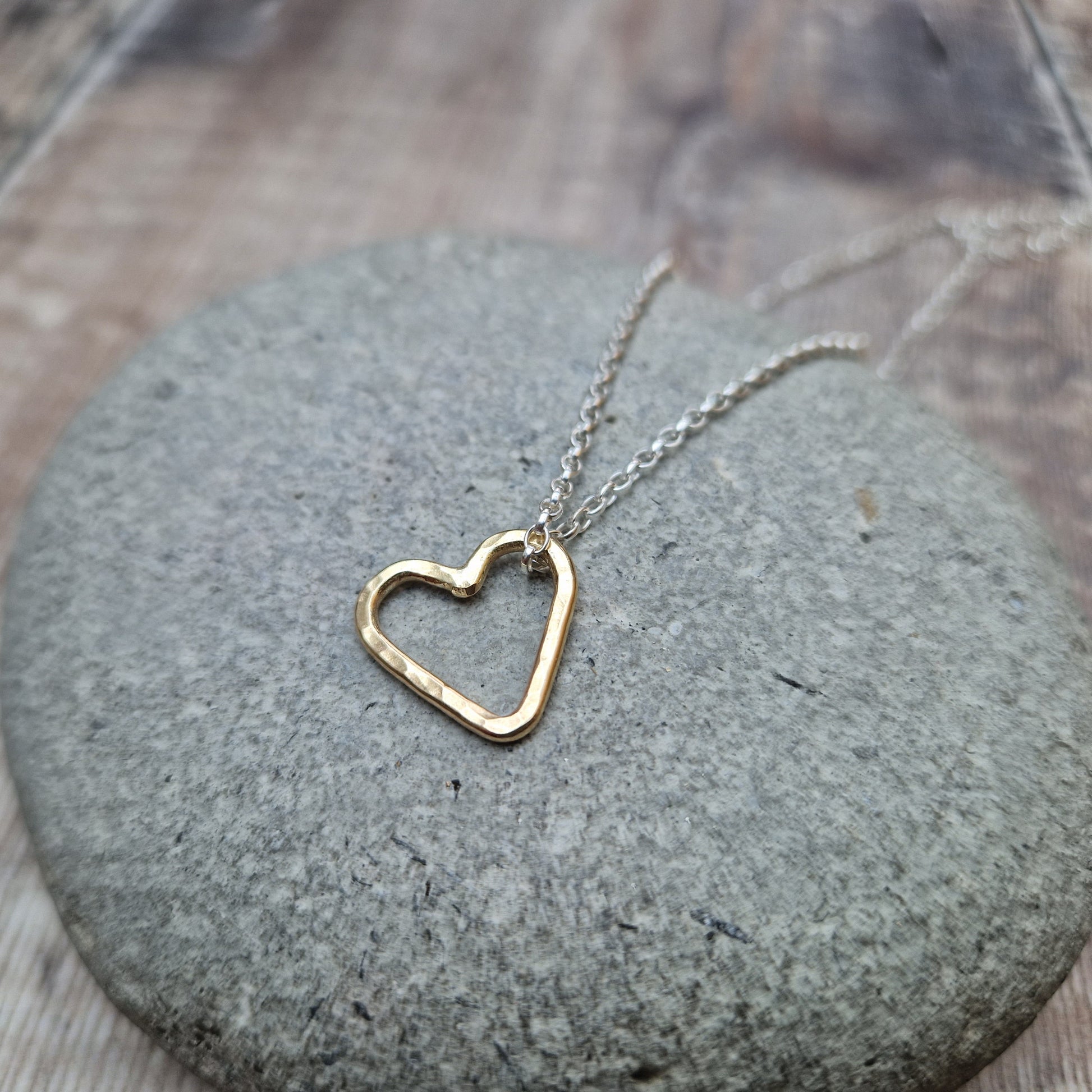 A close-up view of a hammered gold open heart pendant hanging from a silver chain, placed on a smooth stone background.