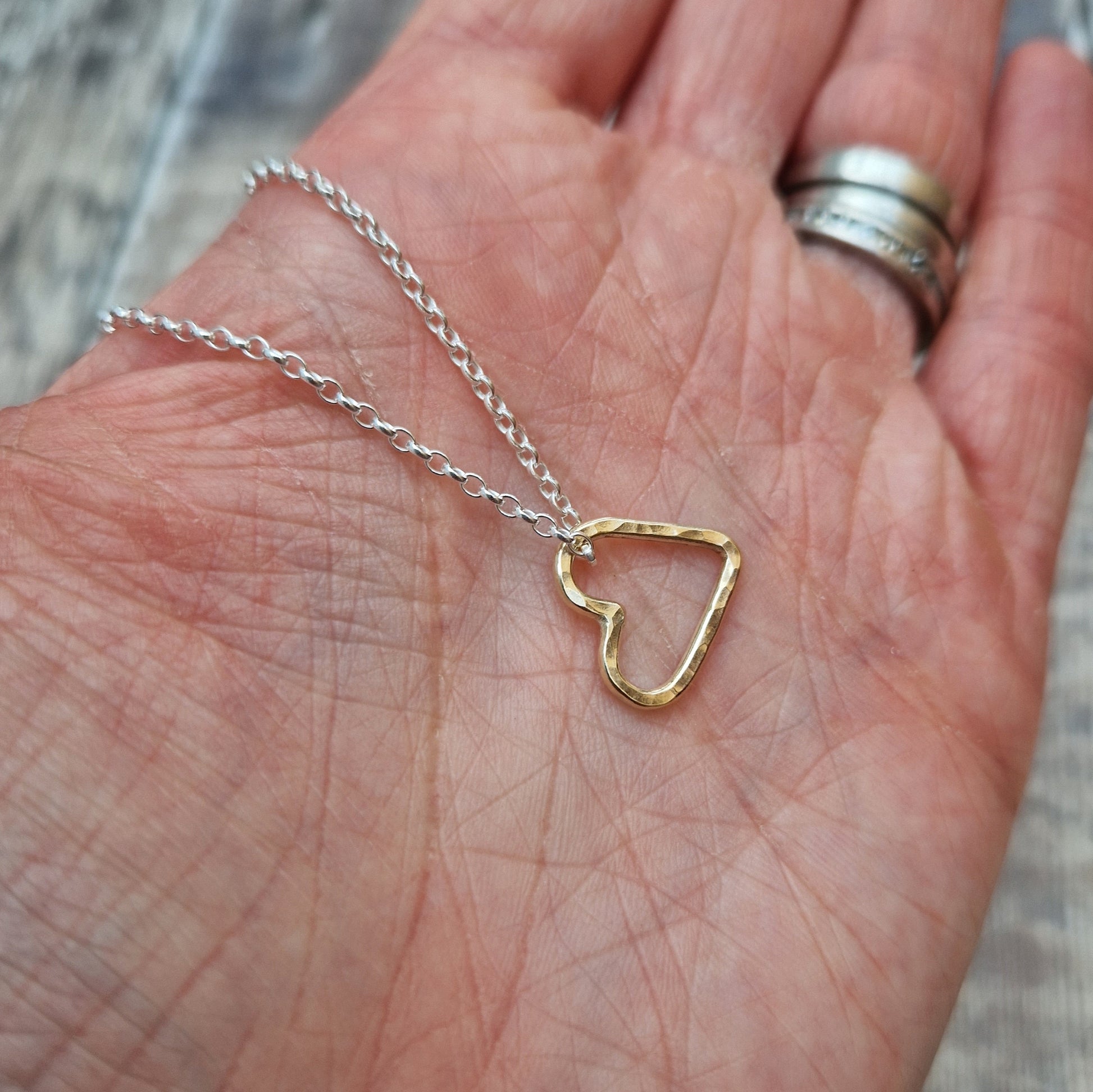 A person holding a delicate silver necklace with a hammered gold open heart pendant in their hand.