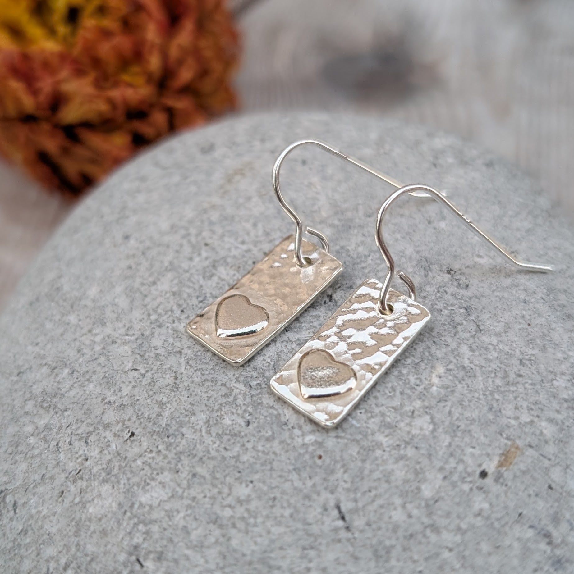 Each earring is a rectangular shape set with a raised heart shape accent onto hammered textured finish. Suspended from silver earring wire.