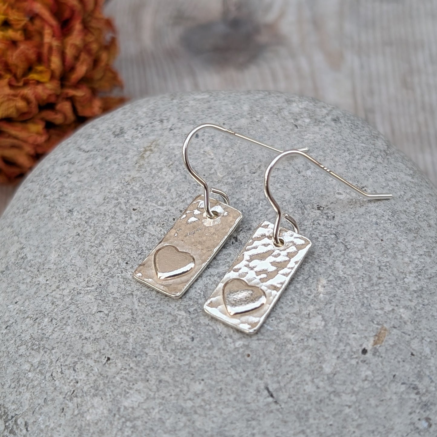 Each earring is a rectangular shape set with a raised heart shape accent onto hammered textured finish. Suspended from silver earring wire.