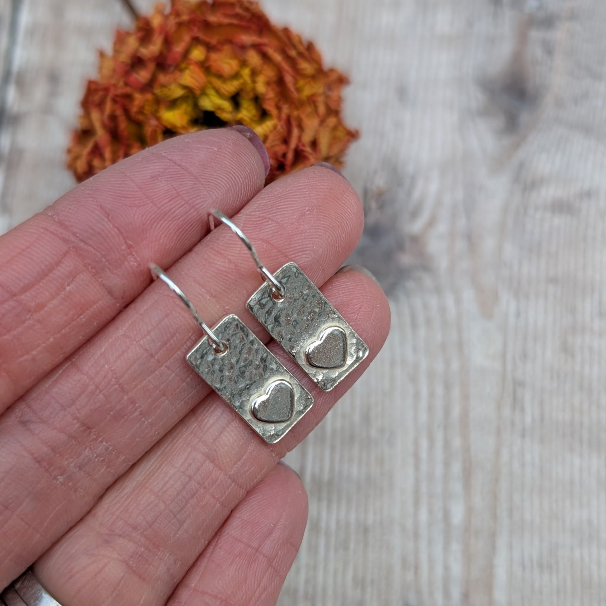 Each earring is a rectangular shape set with a raised heart shape accent onto hammered textured finish. Suspended from silver earring wire.