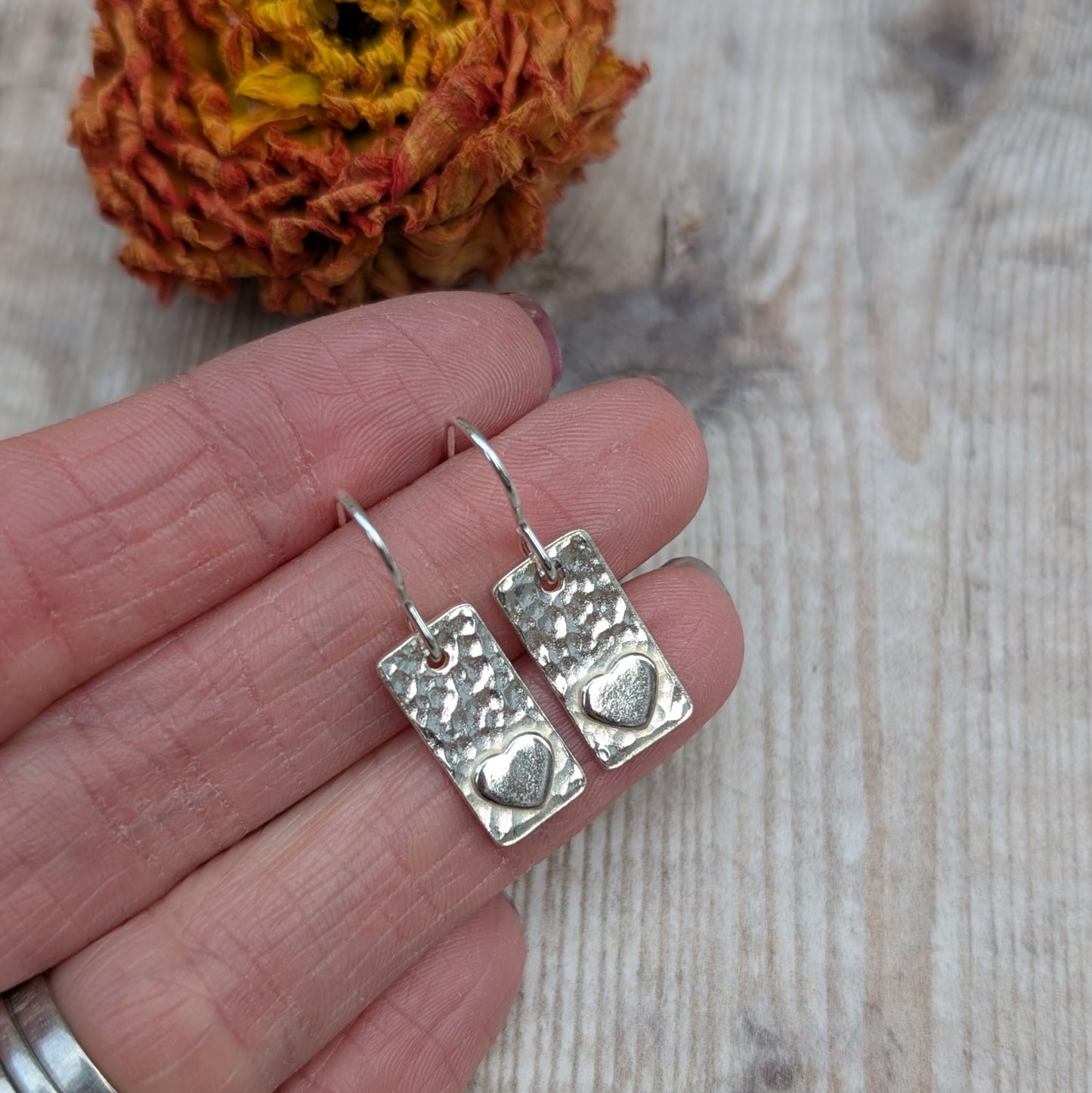 Each earring is a rectangular shape set with a raised heart shape accent onto hammered textured finish. Suspended from silver earring wire.