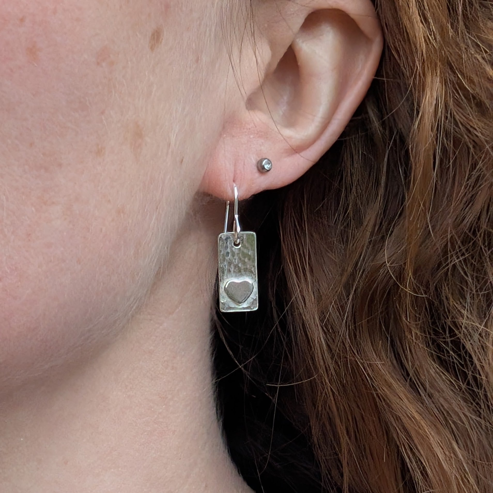 Each earring is a rectangular shape set with a raised heart shape accent onto hammered textured finish. Suspended from silver earring wire.