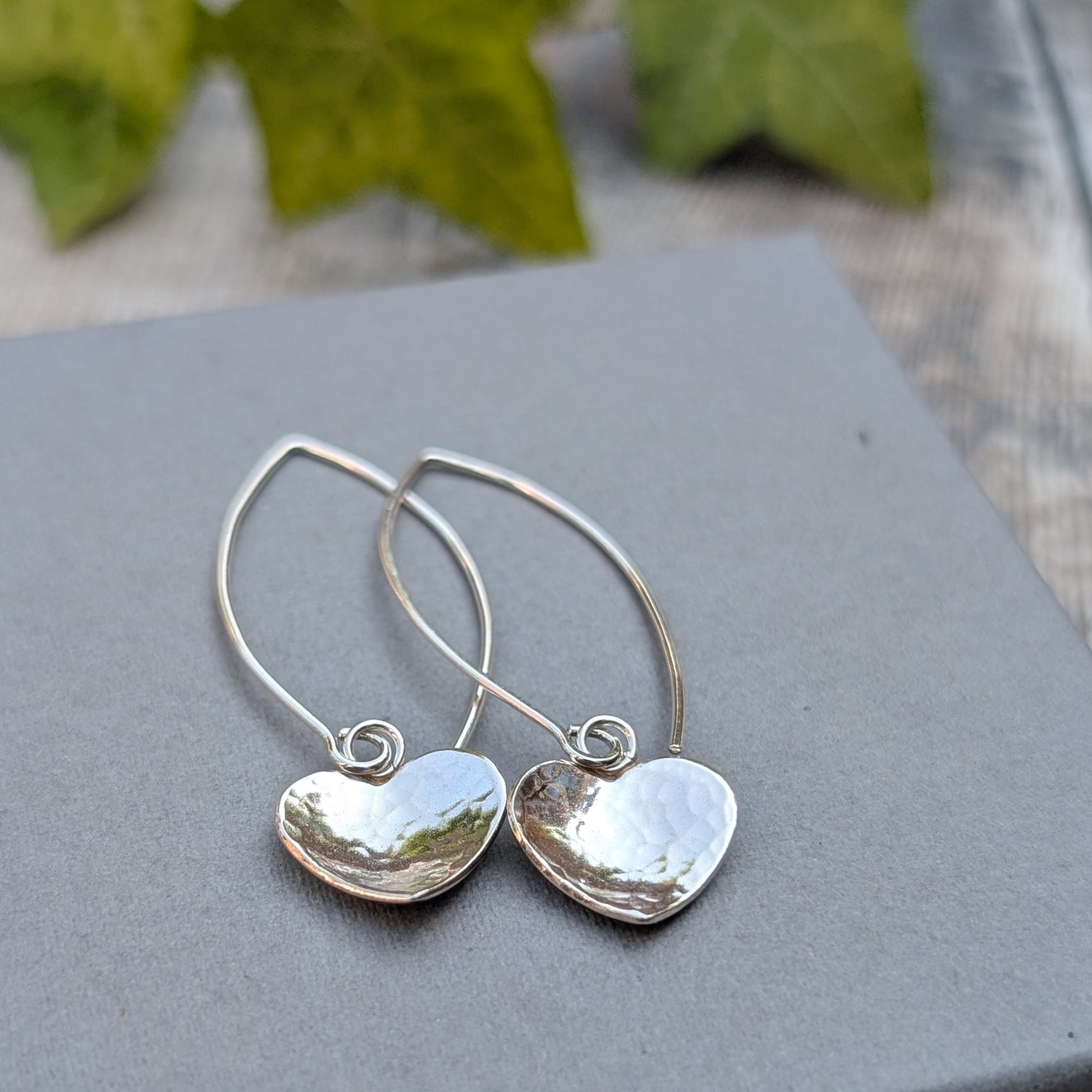 Hammered silver heart long earrings handmade from recycled Sterling Silver, displayed on a grey jewellery box with a natural leafy background.