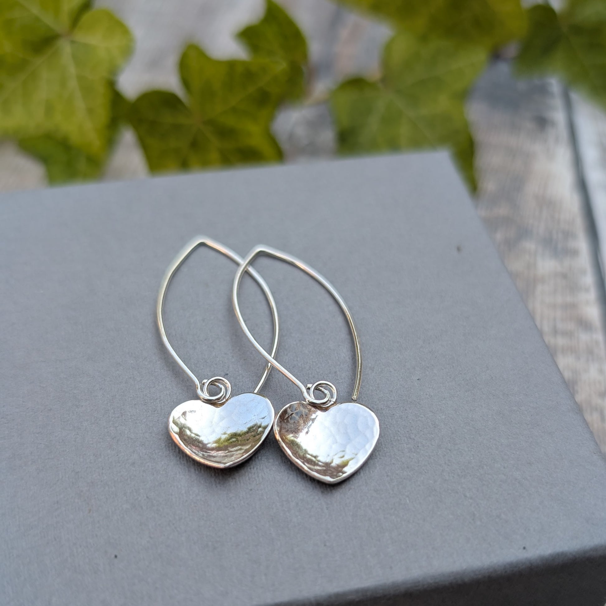 Hammered silver heart long earrings handmade from recycled Sterling Silver, displayed on a grey jewellery box with a leafy background.