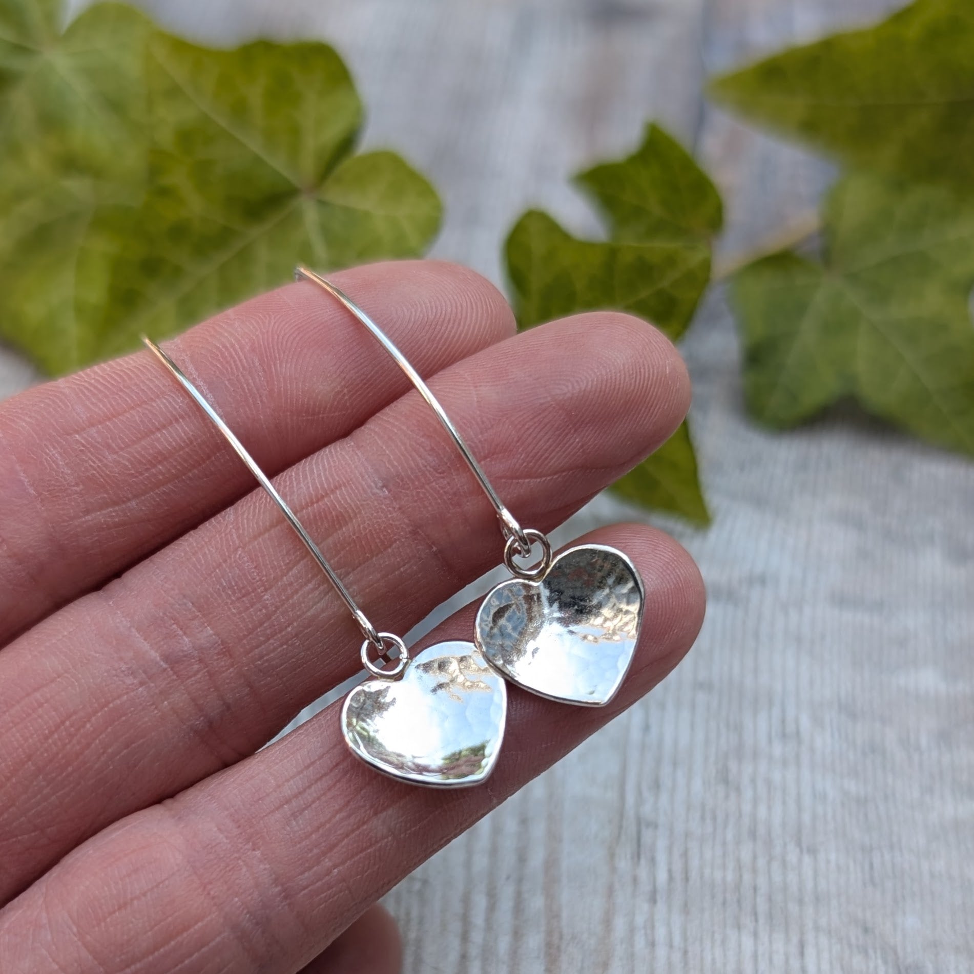 Hammered silver heart long earrings handmade from recycled Sterling Silver, shown resting on fingertips to highlight their size.