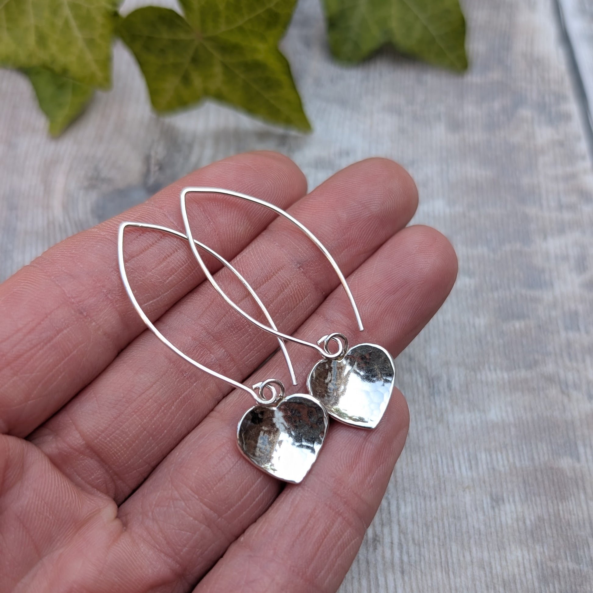Hammered silver heart long earrings handmade from recycled Sterling Silver, displayed on a hand with the long ear wires clearly visible.
