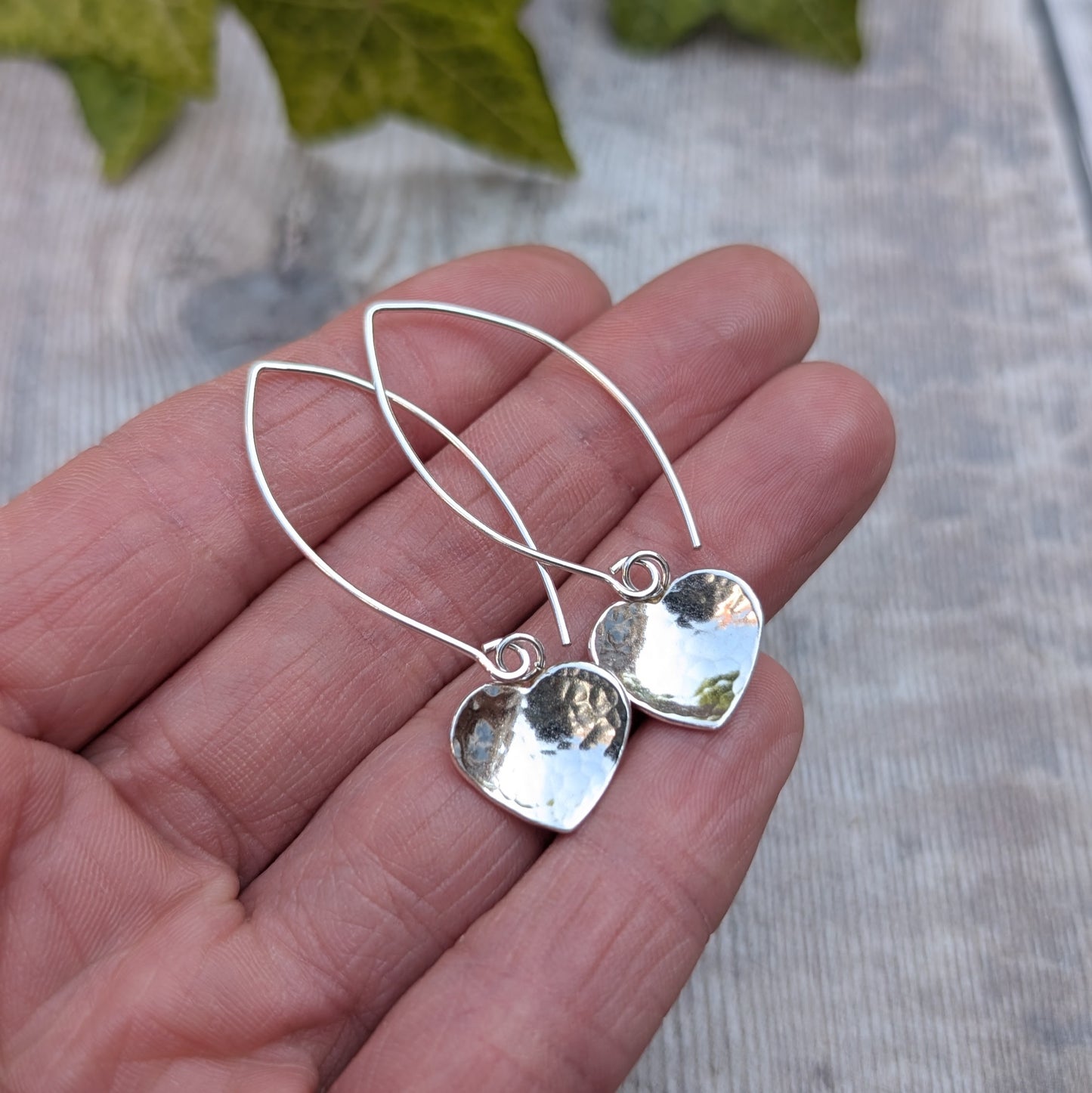 Hammered silver heart long earrings handmade from recycled Sterling Silver, photographed on a hand against a rustic wooden background.