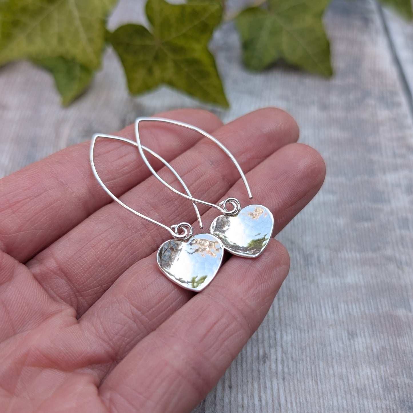 Hammered silver heart long earrings handmade from recycled Sterling Silver, close up on a hand showing the hammered heart detail and polished finish.