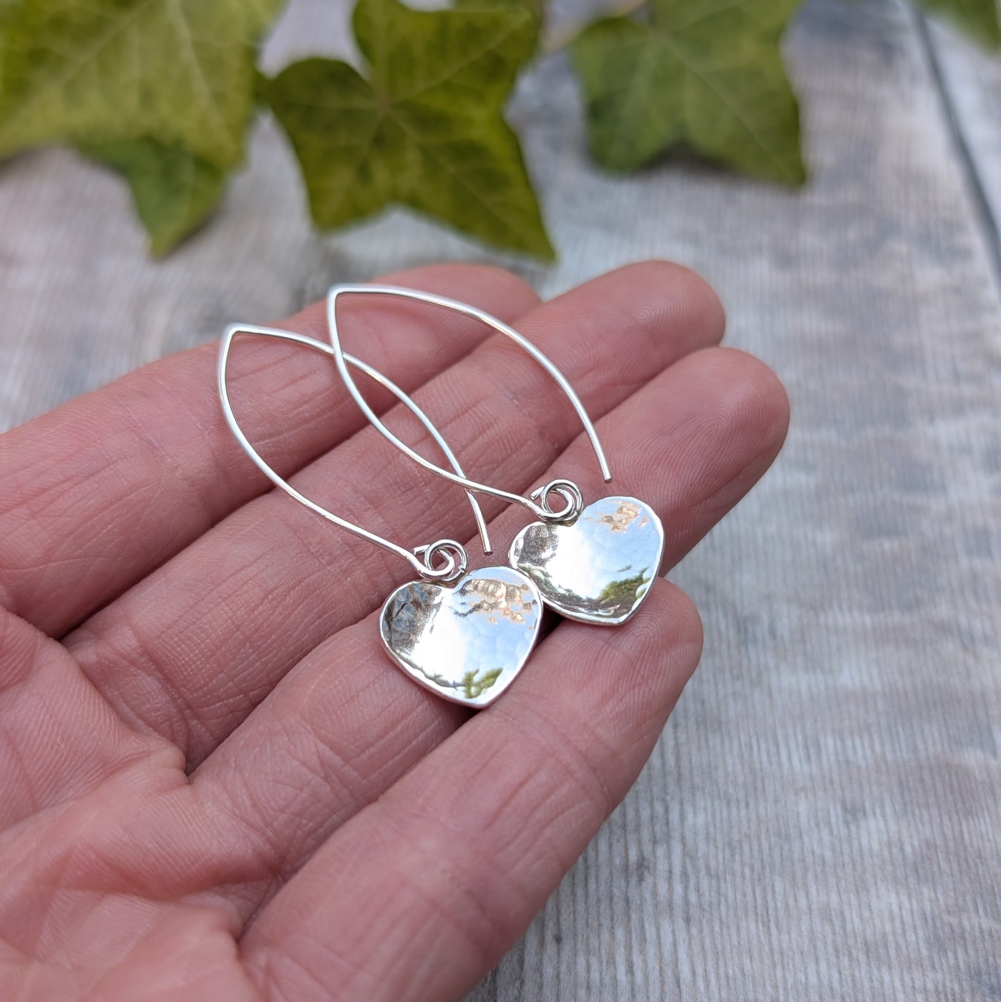 Hammered silver heart long earrings handmade from recycled Sterling Silver, close up on a hand showing the hammered heart detail and polished finish.
