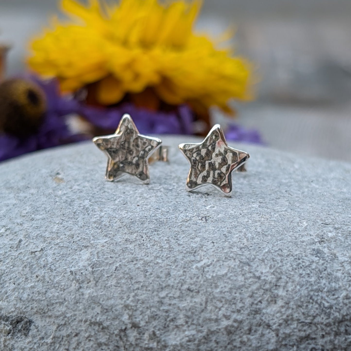 Close-up of a pair of handmade sterling silver star-shaped stud earrings with a hammered texture, resting on a smooth grey stone with vibrant yellow and purple flowers in the background.