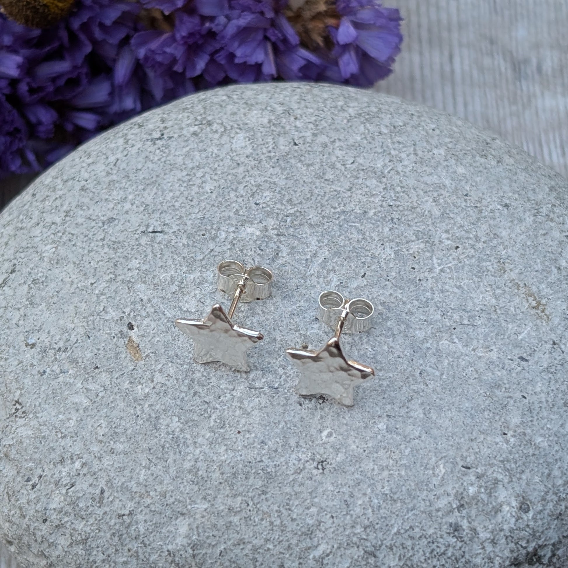 Close-up of a pair of handmade sterling silver star-shaped stud earrings with a hammered texture, resting on a smooth grey stone with vibrant yellow and purple flowers in the background.