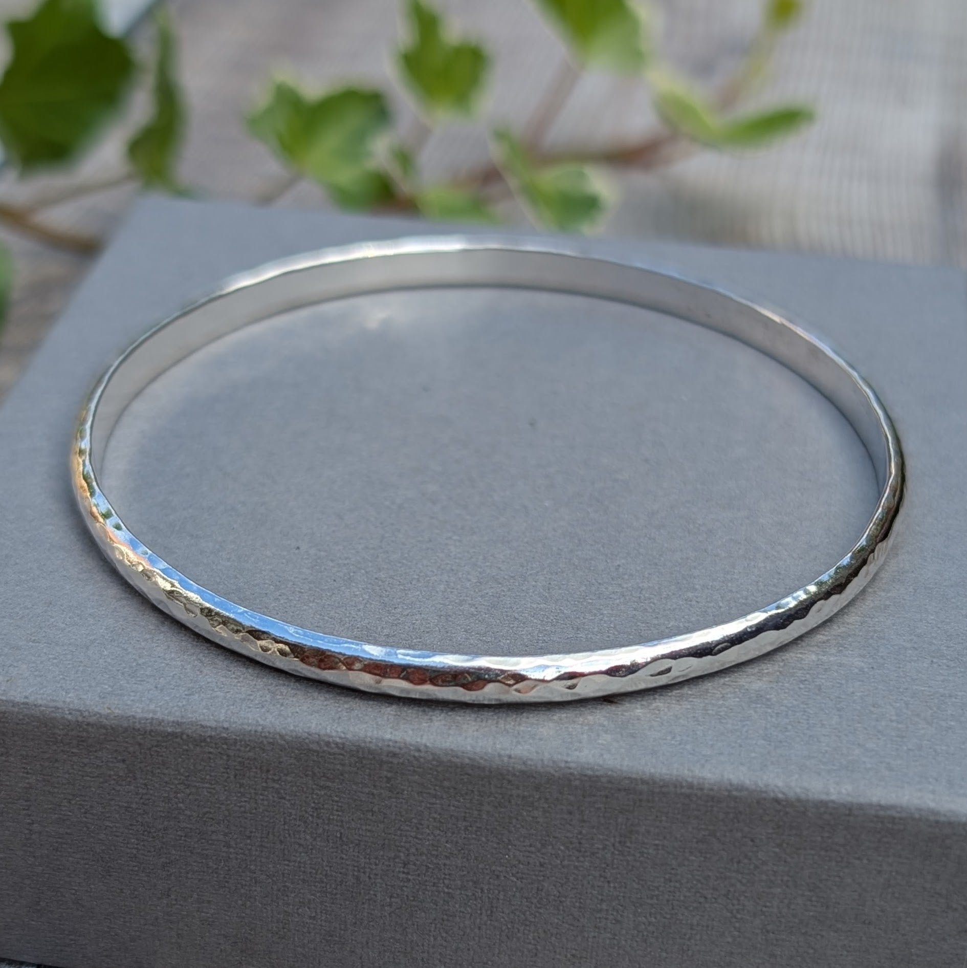 Hammered Sterling Silver D Shape Bangle handmade from recycled Sterling Silver D shaped wire, displayed on a grey jewellery box with a soft green leafy background.