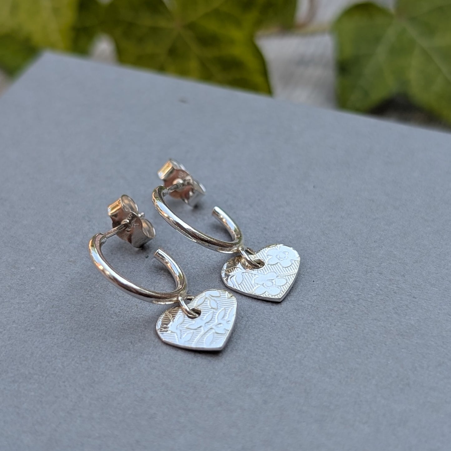Close up of silver heart drop hoop earrings handmade from recycled Sterling Silver with floral textured heart charms displayed on a grey jewellery box.