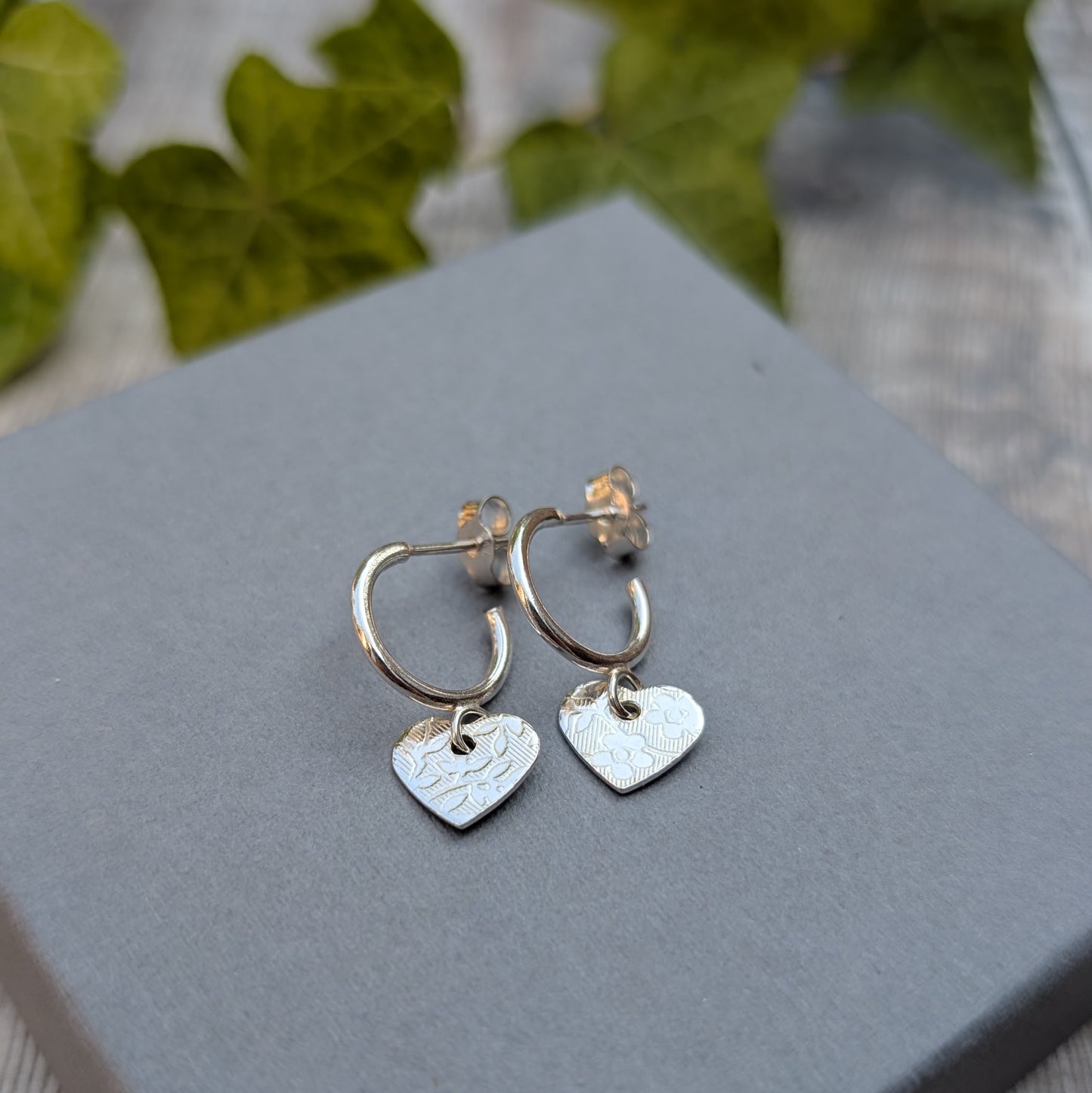 Silver heart drop hoop earrings handmade from recycled Sterling Silver with textured floral heart charms displayed on a grey jewellery box.