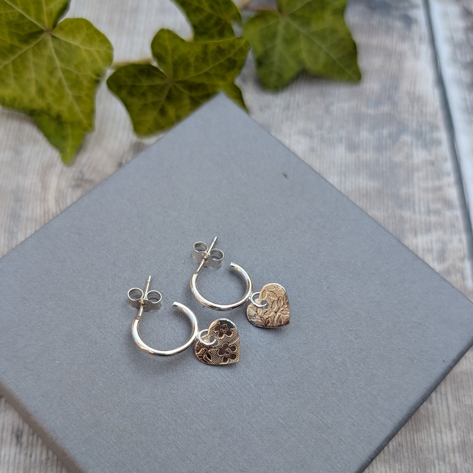 Back view of silver heart drop hoop earrings handmade from recycled Sterling Silver with textured heart charms on a grey jewellery box