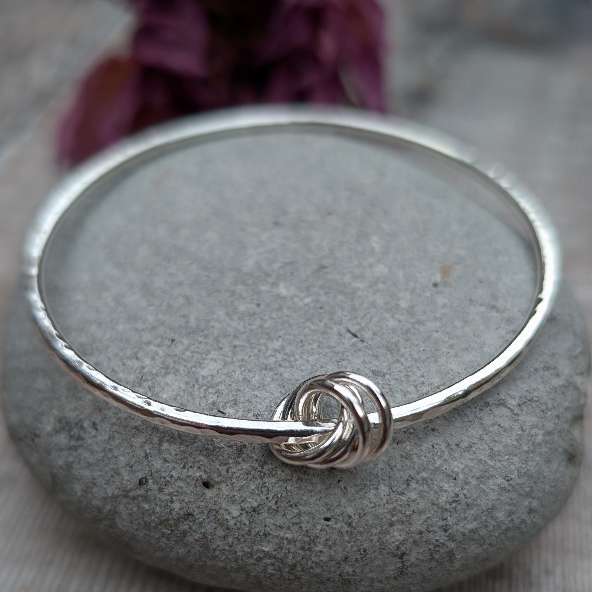 A hammered silver knot bangle resting on a smooth grey stone with a decorative purple flower in the background, highlighting the intricate knot detail.