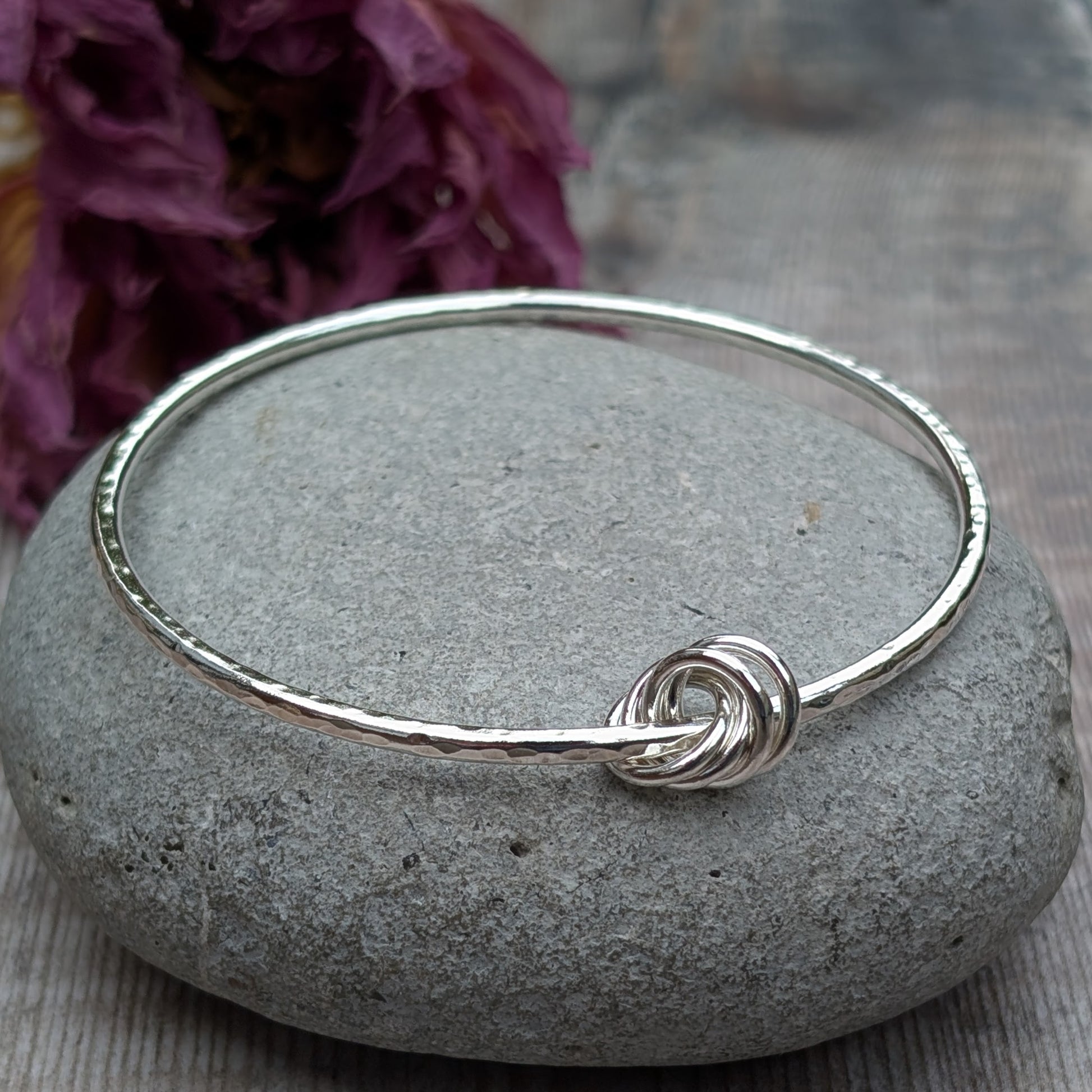 A hammered silver knot bangle resting on a smooth grey stone with a decorative purple flower in the background, highlighting the intricate knot detail.