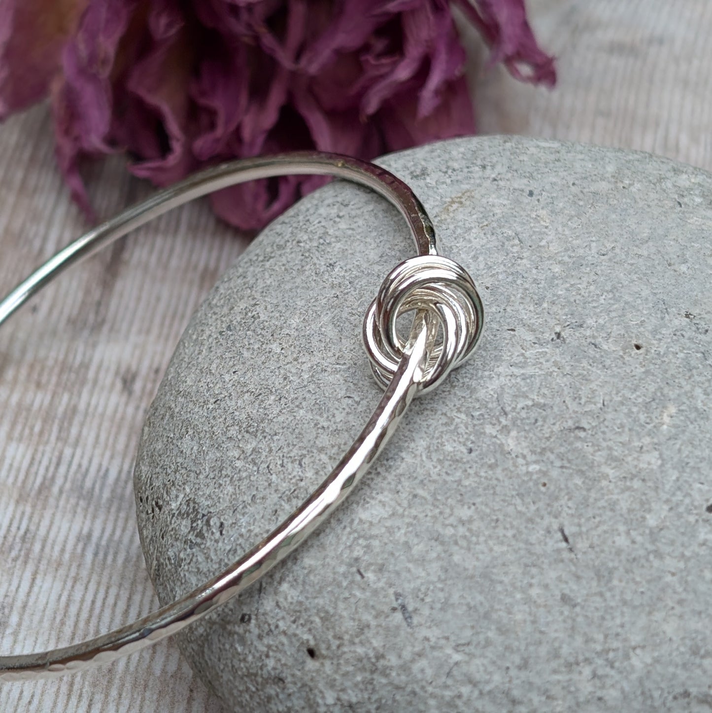 A hammered silver knot bangle resting on a smooth grey stone with a decorative purple flower in the background, highlighting the intricate knot detail.