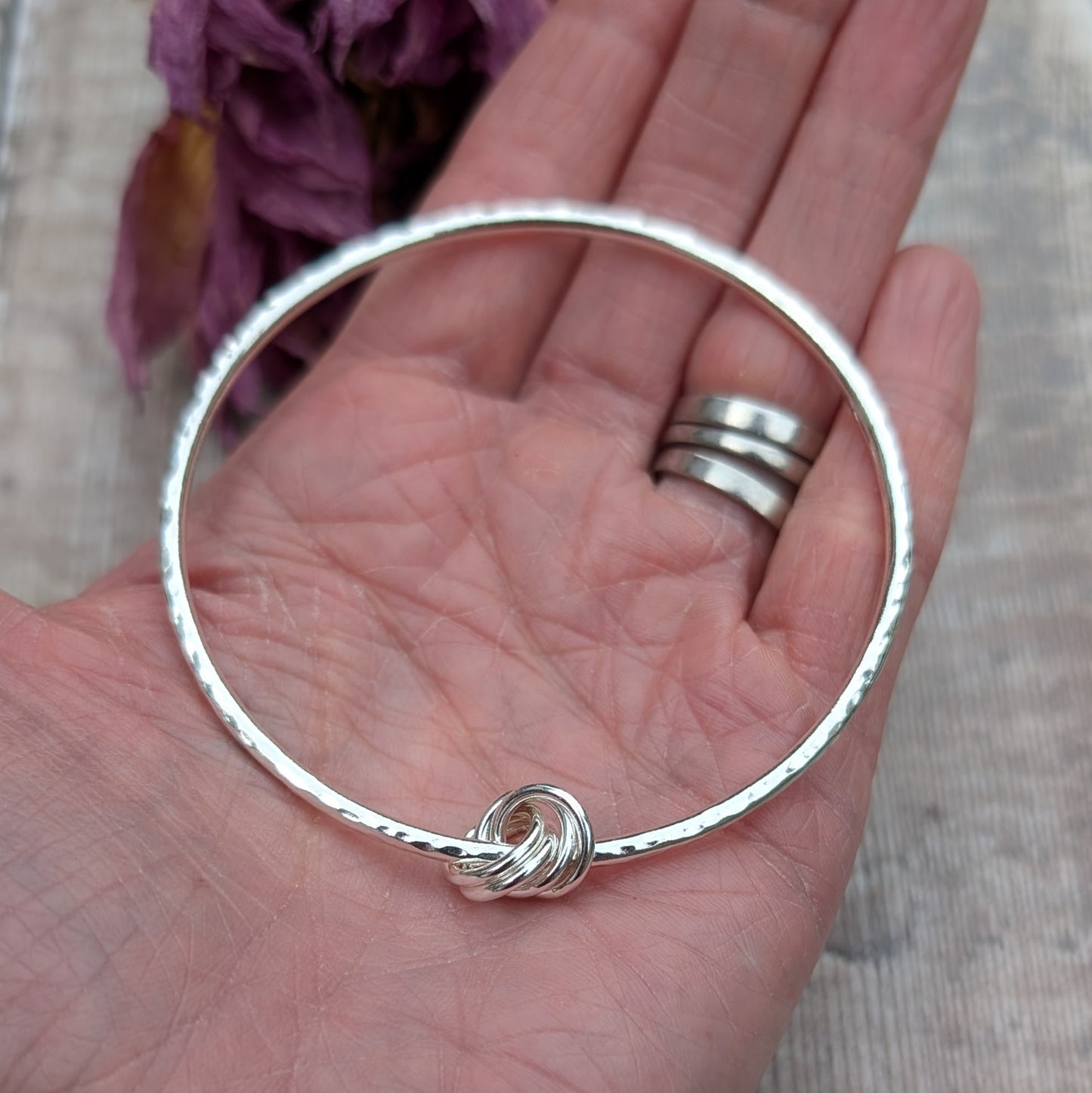 A hammered silver knot bangle being held in a hand, showing its delicate size and craftsmanship, with a blurred purple flower in the background.