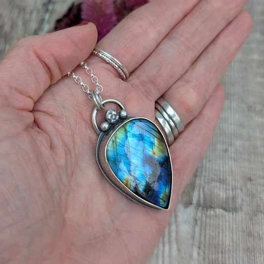 A close-up of a hand holding the labradorite pendant, showcasing the gemstone’s iridescent blue and gold flashes. The pendant is set in a recycled sterling silver bezel with a decorative silver bead detail at the top.