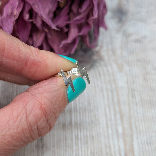 A hand holding a small pair of silver lightning bolt stud earrings, with a close-up view showing their sharp design and matte finish.