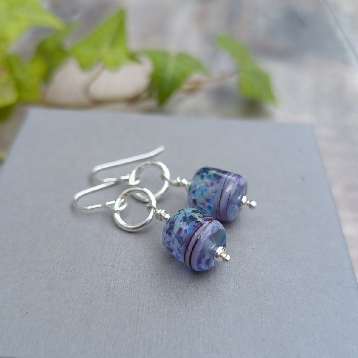 Close up of Lilac Watercolour Lampwork Glass and Silver Circle Earrings on a grey box, showing the purple, blue and green handmade Lampwork glass beads.