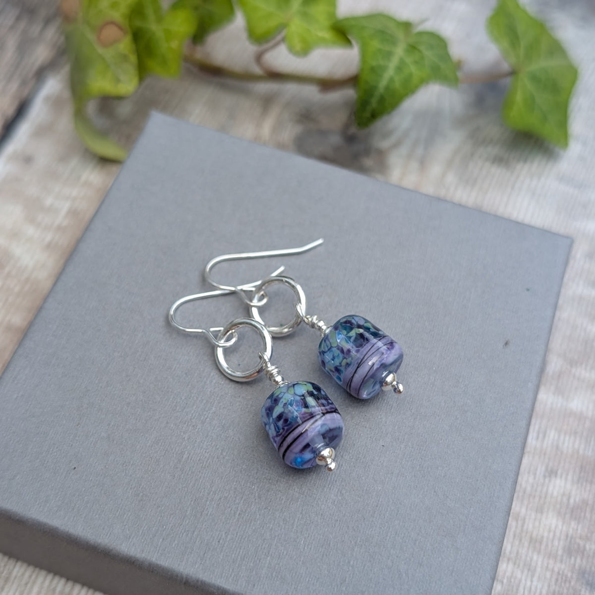 Lilac Watercolour Lampwork Glass and Silver Circle Earrings handmade from recycled Sterling Silver with lilac, blue and green glass beads, displayed on a grey box.