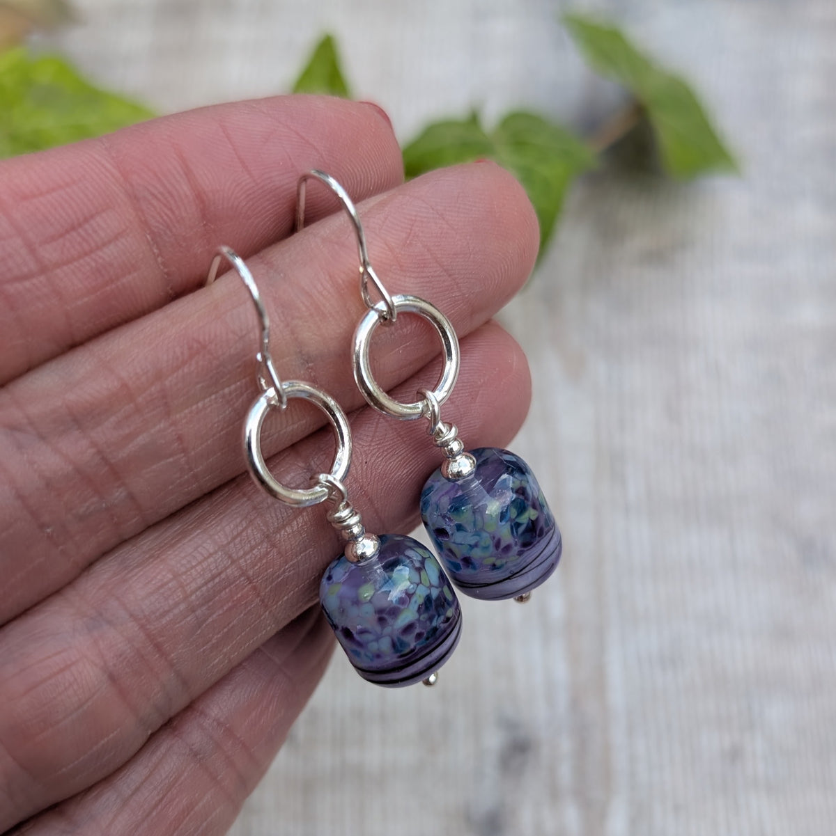 Lilac Watercolour Lampwork Glass and Silver Circle Earrings held in a hand, showing the lilac, blue and green pattern of the handmade Lampwork glass beads.