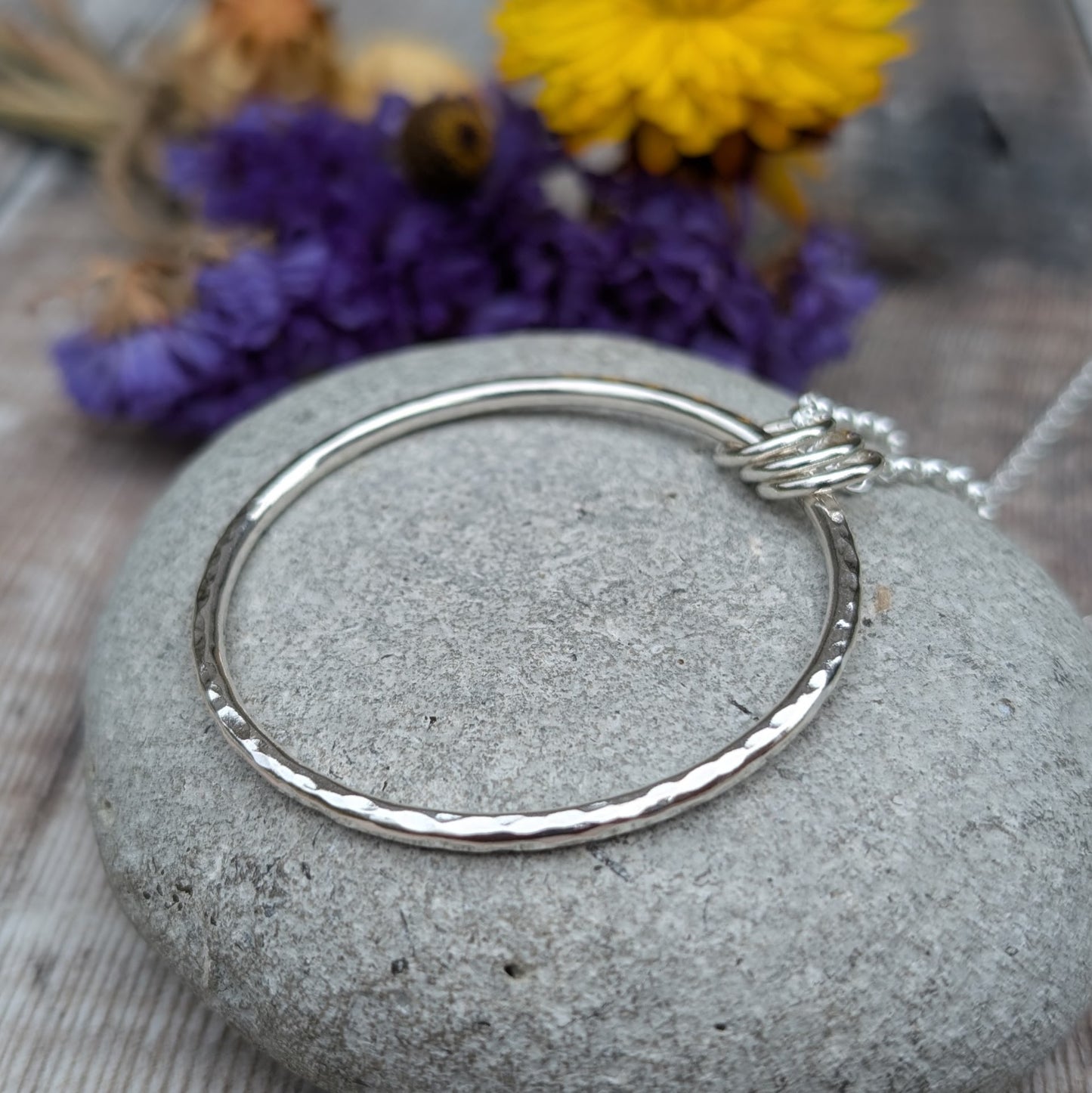 A silver hammered circle pendant with three silver loops resting on a smooth grey stone, with blurred yellow and purple dried flowers in the background.