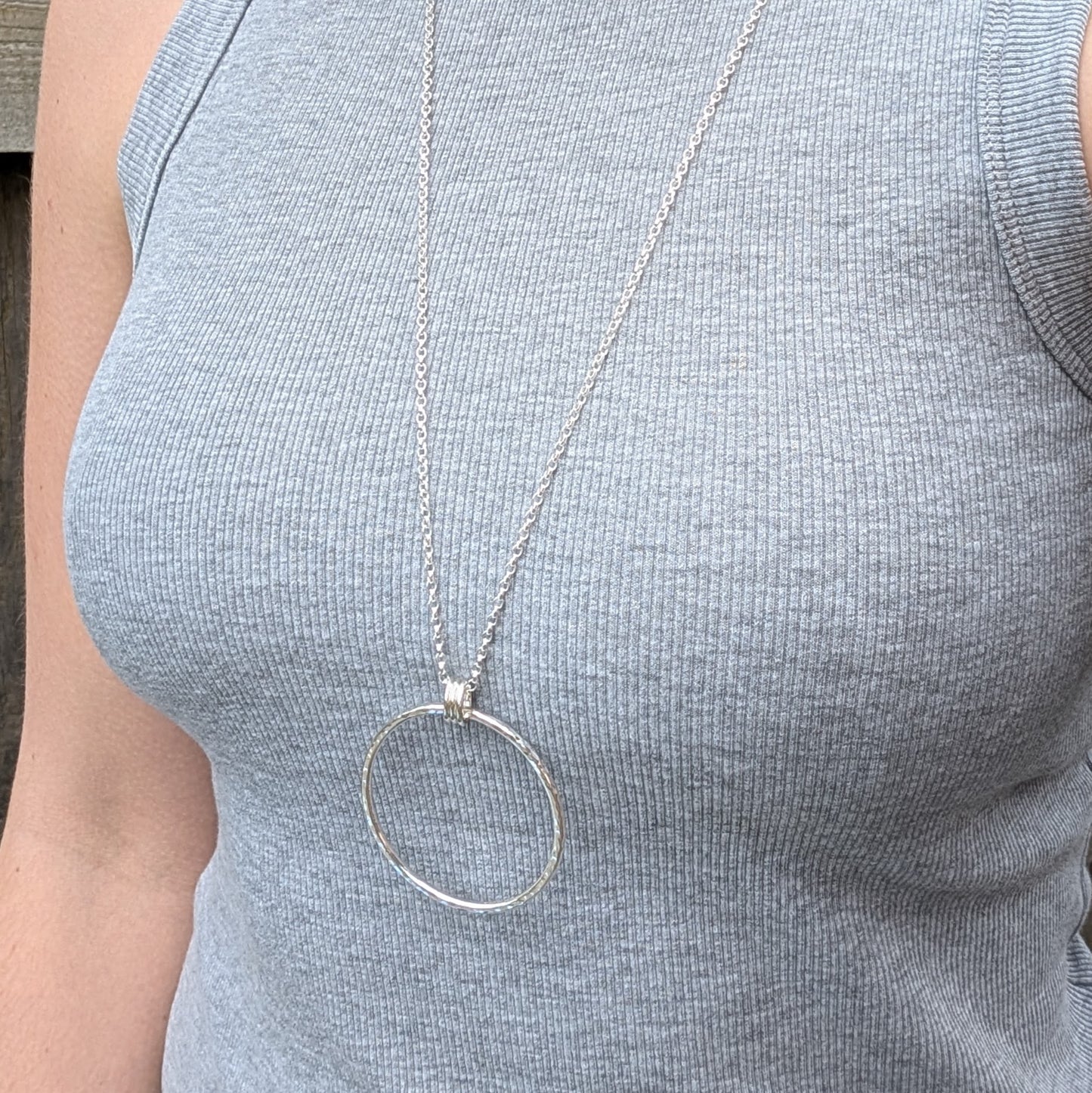 A person wearing a long silver necklace with a large hammered circle pendant resting on the chest, over a ribbed grey sleeveless top.