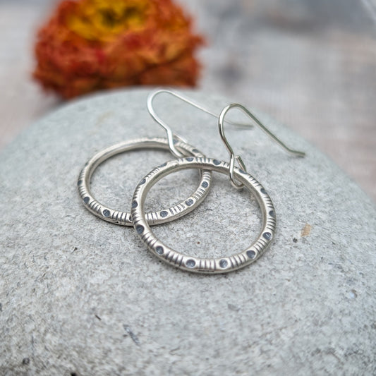 Each earring is a sterling silver hoop stamped with a repeating pattern of moon as a dot and four lines, ‘lunar lines’ pattern, suspended from silver earring wire.